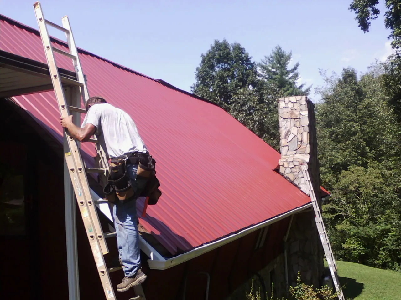 Roofer on a ladder beside a red metal roof and stone chimney — Soffit & Fascia Repair by Roofing Brothers in Breezy Point